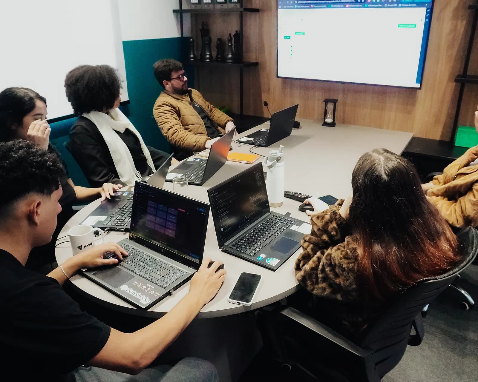 Reunião na sala de reunião da Hype Mídia, com seis pessoas à mesa acompanhando uma apresentação na TV